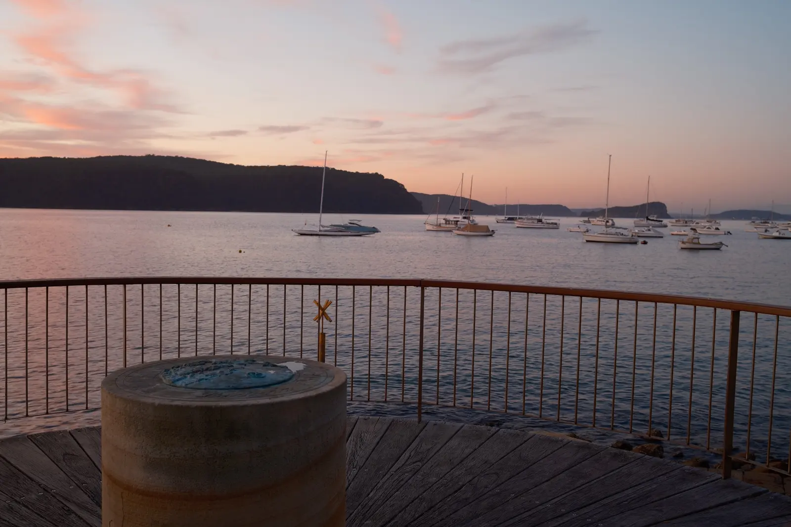 Yachts moored on calm water at dusk, seen over the railing of a curved timber lookout with a bronze map on a sandstone plinth